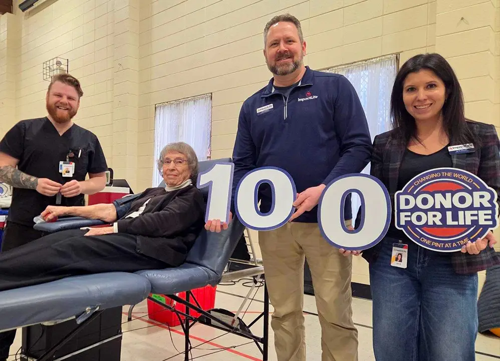 Elderly woman donating blood on a recliner while three volunteers stand beside her, holding a large '100' sign and a 'Donor for Life' badge at a blood drive location.